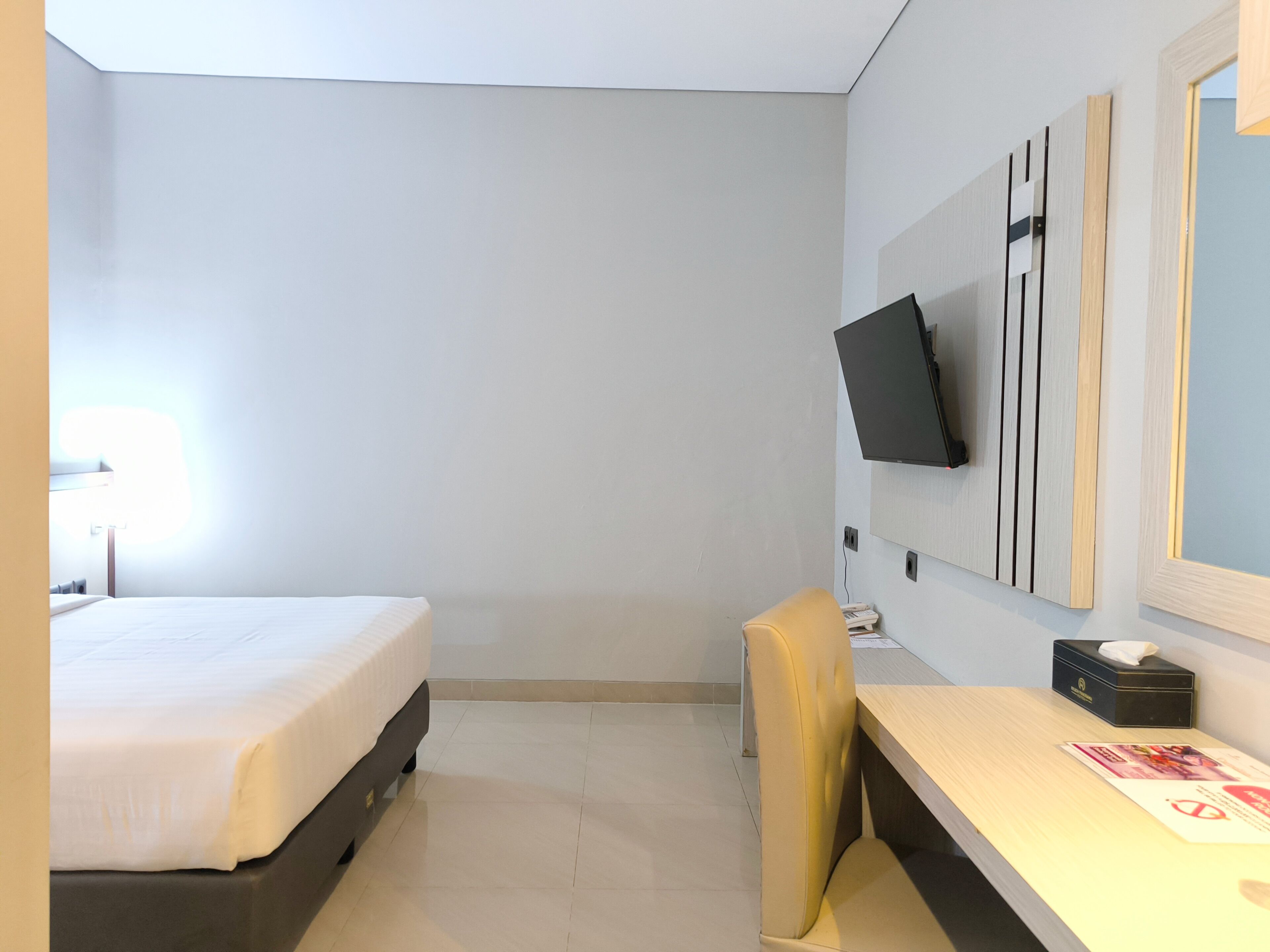 A hotel room with a bed, a desk, a chair, a TV mounted on the wall, and a mirror.