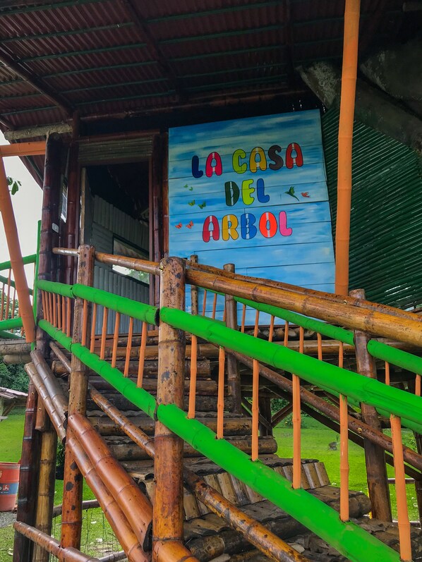 A wooden staircase with green railings leading to a covered area with a sign reading "LA CASA DEL ARBO".