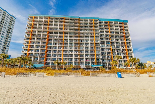 A multi-story hotel with balconies, located near a sandy beach.