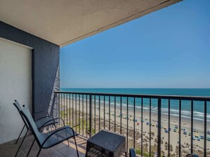 Un balcon donnant sur la plage, des chaises en métal, une petite table et une rambarde.