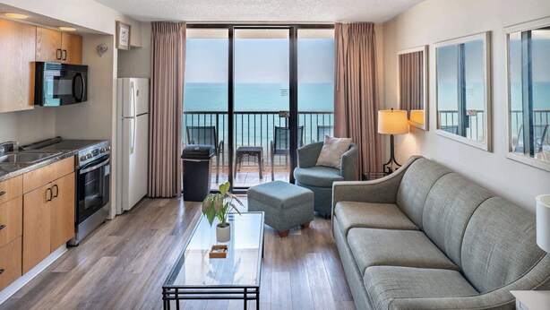 Een moderne hotelkamer met een balkon, een kitchenette, een bank en een glazen salontafel.