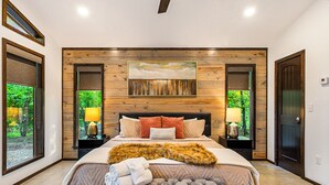 A bedroom with a large bed, wooden panel wall, two bedside lamps, and a view of greenery through the windows.