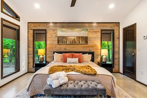 A bedroom with a large bed, wooden panel wall, two bedside lamps, and a view of greenery through the windows.