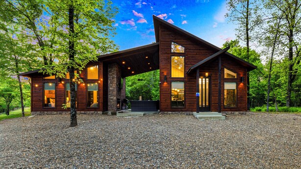 A modern log cabin with a stone foundation, large windows, and a gravel driveway.