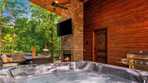 A hot tub in a log cabin with a stone fireplace, outdoor seating, and a television mounted on the wall.