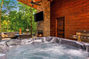 A hot tub in a log cabin with a stone fireplace, outdoor seating, and a television mounted on the wall.