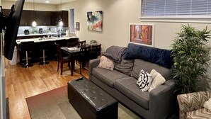 Una sala moderna con un sofá gris, un reposapiés negro y un comedor con cocina al fondo.