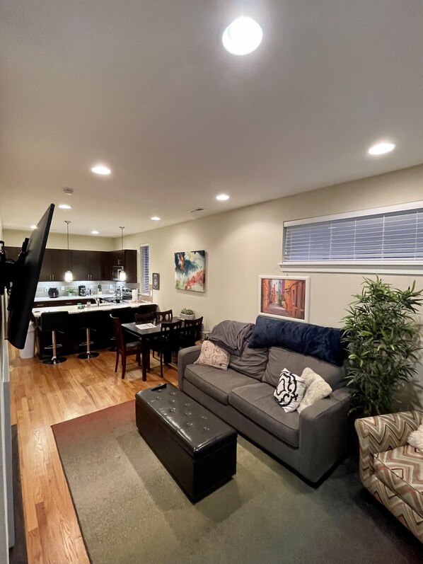 A modern living room with a grey sofa, a black ottoman, and a dining area with a kitchen in the background.