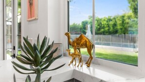 A window with a view of greenery, a golden camel figurine, and a plant on a white ledge. - Walkable Historic Phoenix Casita + Private Pool (Phoenix)