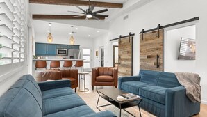 A modern living room with a blue sofa, a leather armchair, and a coffee table. There is a kitchen in the background with teal cabinets and a hanging light fixture. - Walkable Historic Phoenix Casita + Private Pool (Phoenix)