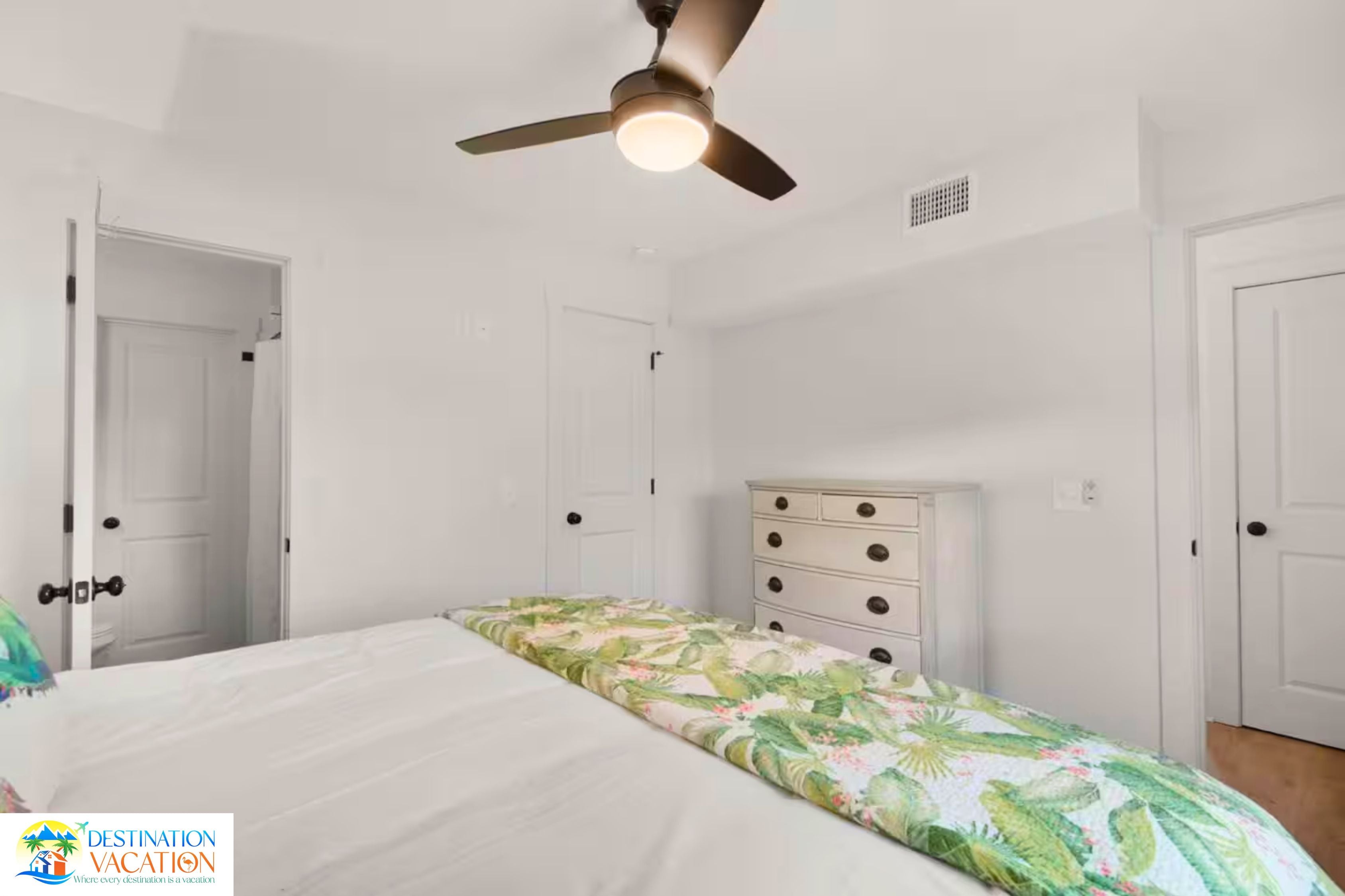 A bedroom with a bed, a dresser, a ceiling fan, and white walls.