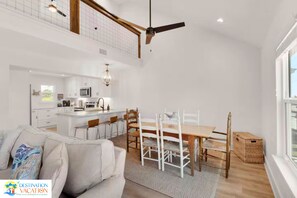 Een ruime woonkamer met een hoge plafond, een eettafel met stoelen, een keuken met moderne apparatuur en een groot raam dat voor natuurlijk licht zorgt.