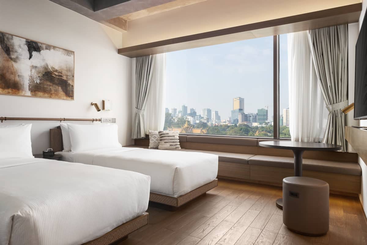 Ein modernes Hotelzimmer mit zwei Betten, einem großen Fenster mit Vorhängen, einem runden Tisch und Blick auf eine Stadtlandschaft.