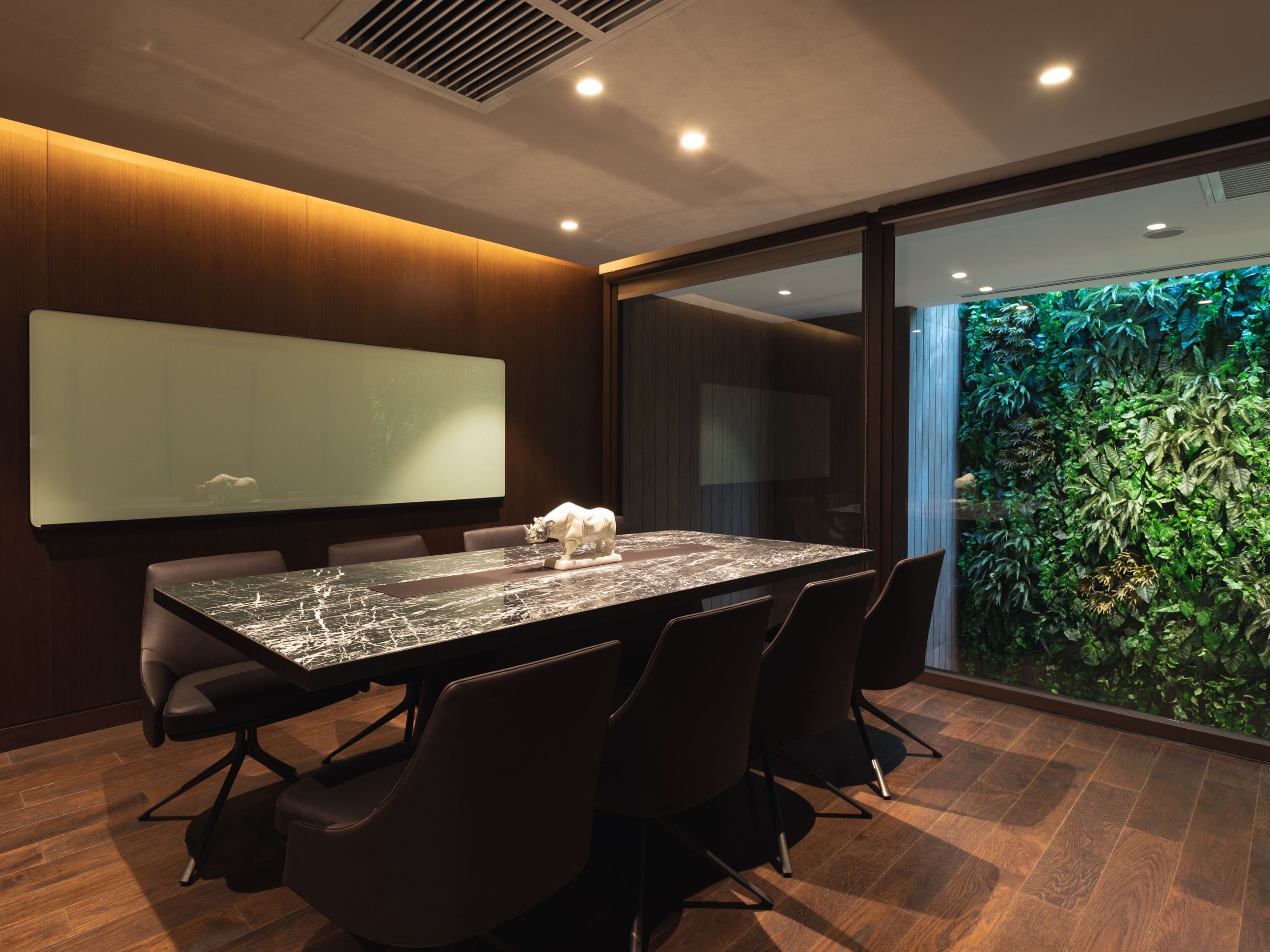 A conference room with a large table, chairs, a projector screen, and a view of a green wall with plants.