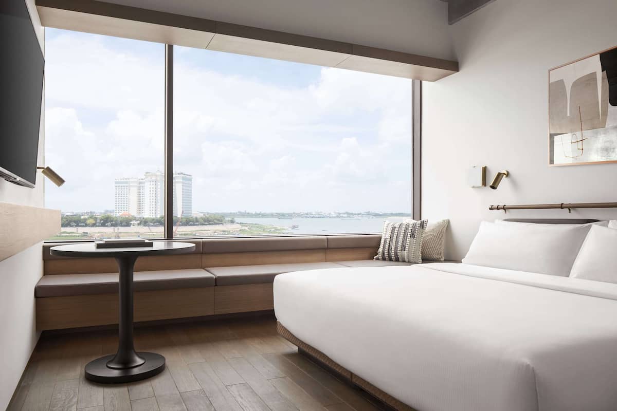 Ein modernes Hotelzimmer mit großem Fenster, einem Bett, einem kleinen runden Tisch und einer Bank.