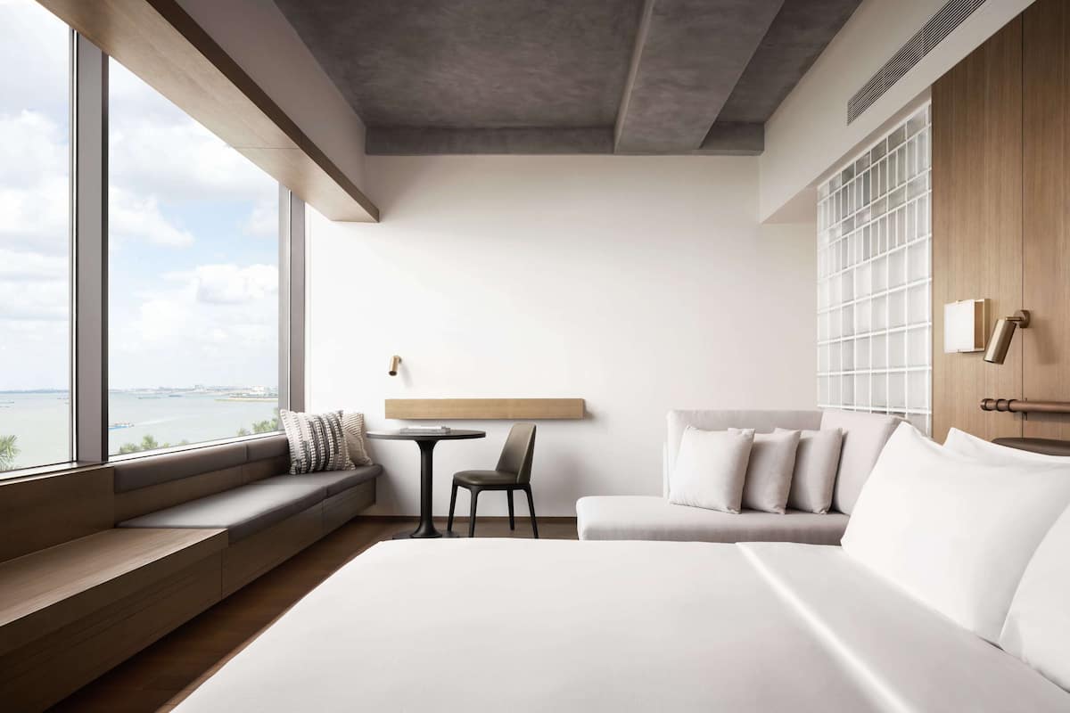 Ein modernes Hotelzimmer mit einem großen Bett, einer Sitzecke mit Tisch und Stühlen sowie Blick auf die Stadt durch große Fenster.