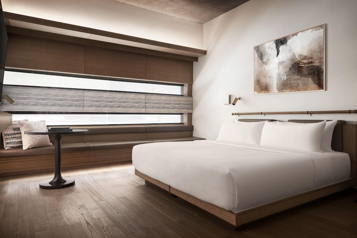 Ein modernes Schlafzimmer mit einem großen Bett, einem kleinen Tisch, einem Fenster mit Jalousien und einem Bild an der Wand.