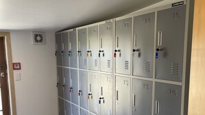 Une rangée de casiers métalliques gris, chacun doté d’un cadenas numéroté, un détecteur d’incendie et une porte en bois.