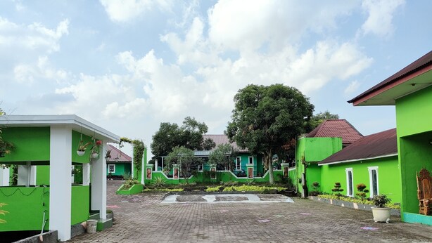 A courtyard with green buildings, a paved area with a red arrow, and a large tree in the center. - Hotel Bukit Sentabi Penembahen (Medan)