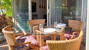 A glass-top dining table with wicker chairs and purple cushions, set on a tiled patio with a view of greenery through a sliding glass door.