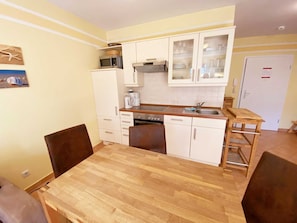 A compact kitchen with white cabinets, a microwave, a stove, and a sink. There is a wooden table with chairs and a small wooden shelf.