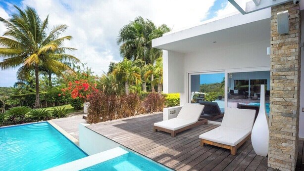 Un espace piscine extérieur moderne, doté de chaises longues, d’une terrasse en bois et offrant une vue sur une maison avec un mur en pierre.