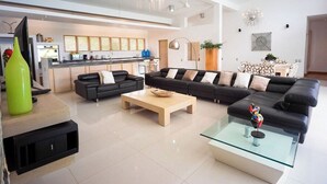 Un salon moderne avec un canapé noir, une table basse en bois et une table basse en verre. On aperçoit une cuisine à l’arrière-plan, avec une fenêtre et une plante.