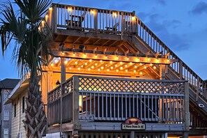 Eine Holzterrasse mit Lichterketten, einem Schild mit der Aufschrift 'A Wave From It All' und einer Palme.