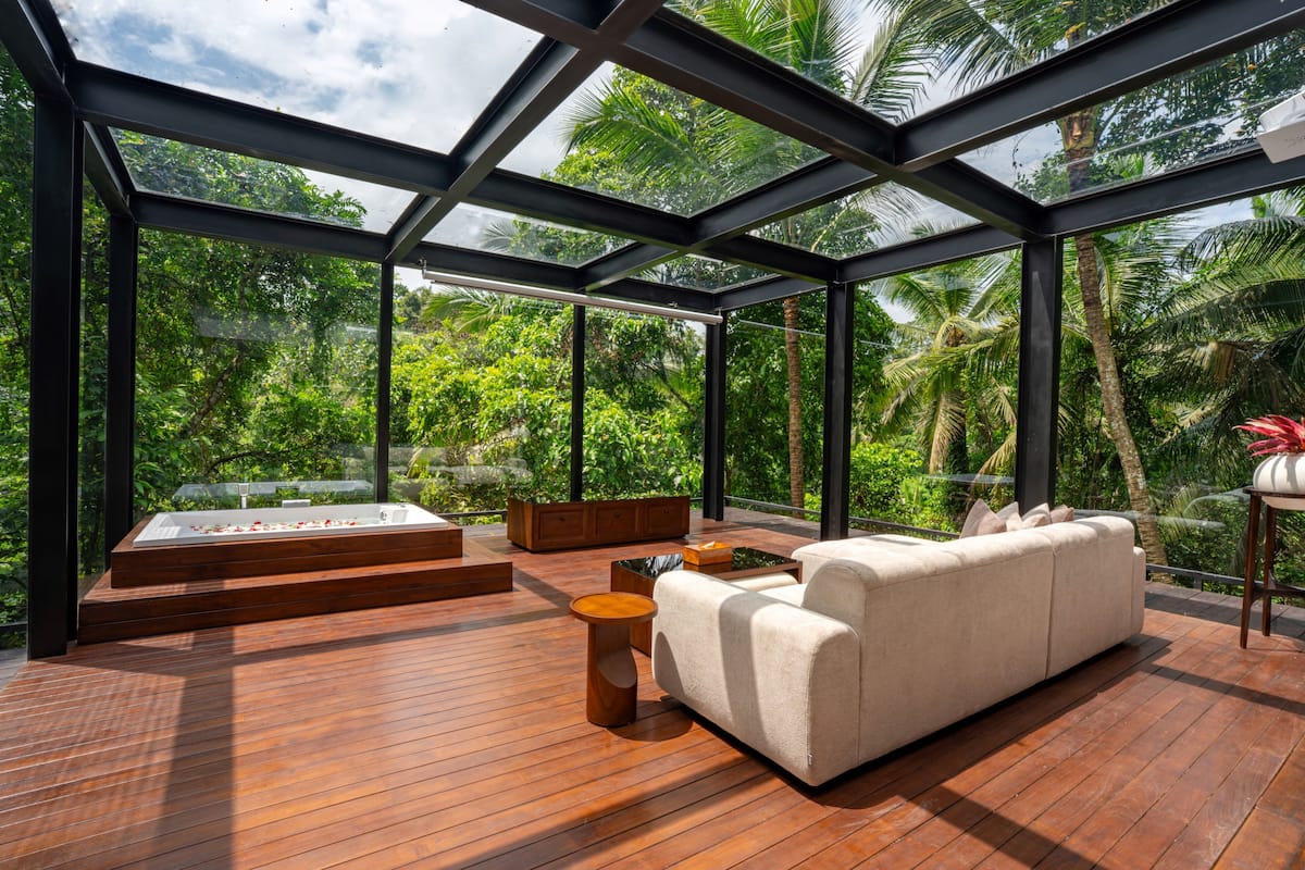 A modern outdoor lounge with a glass ceiling, wooden flooring, a sofa, a side table, and a hot tub.