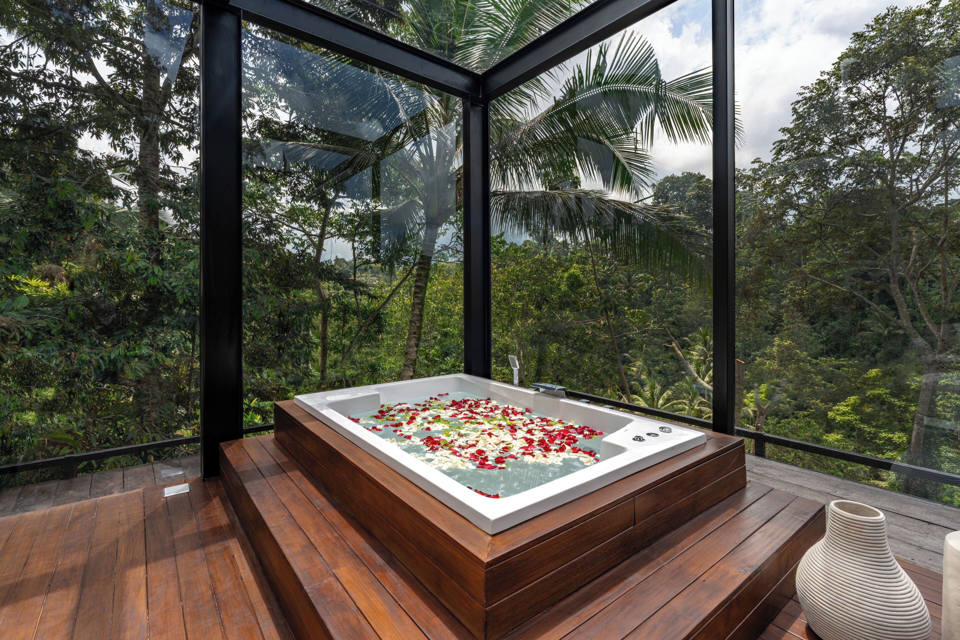 A hot tub with flower petals on a wooden deck, surrounded by glass walls with a forest view.