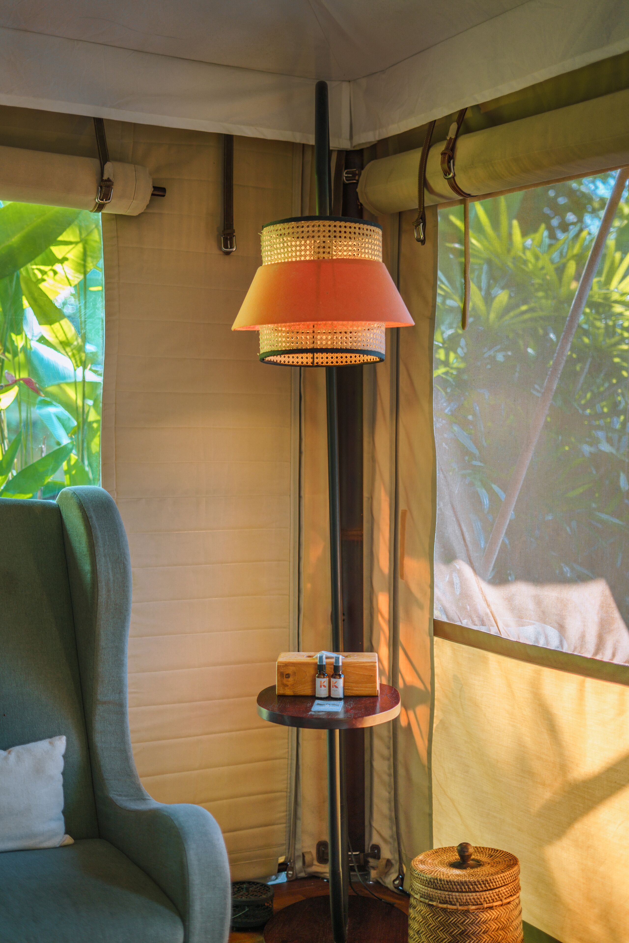 Ein Zimmer mit einem grünen Sessel, einer hohen Stehlampe, einem kleinen Tisch mit Flaschen und einem gewebten Korb.