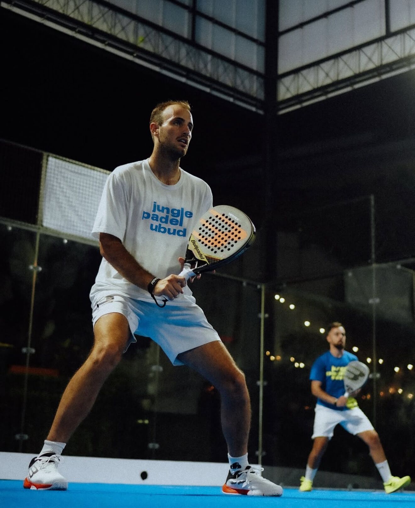 Jugador de tenis con camisa y pantalones cortos en una cancha cubierta, sosteniendo una raqueta.