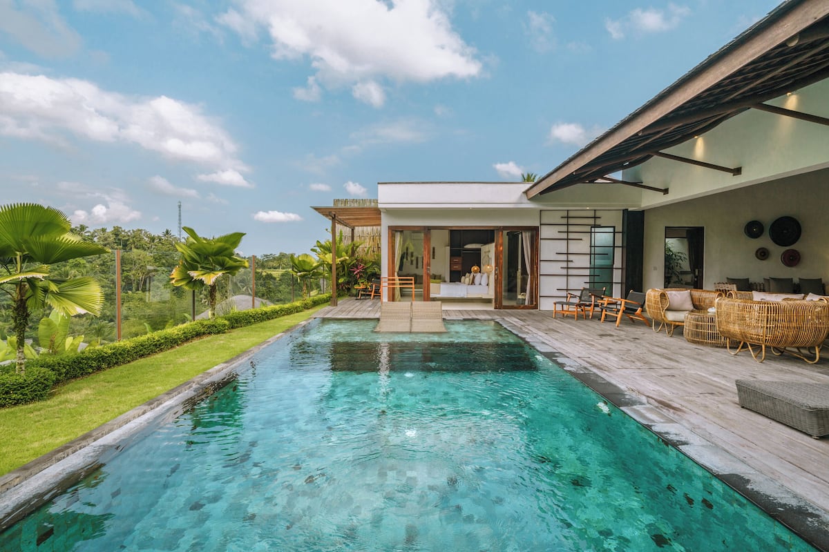 A swimming pool with a wooden deck, wicker furniture, and a modern house in the background.