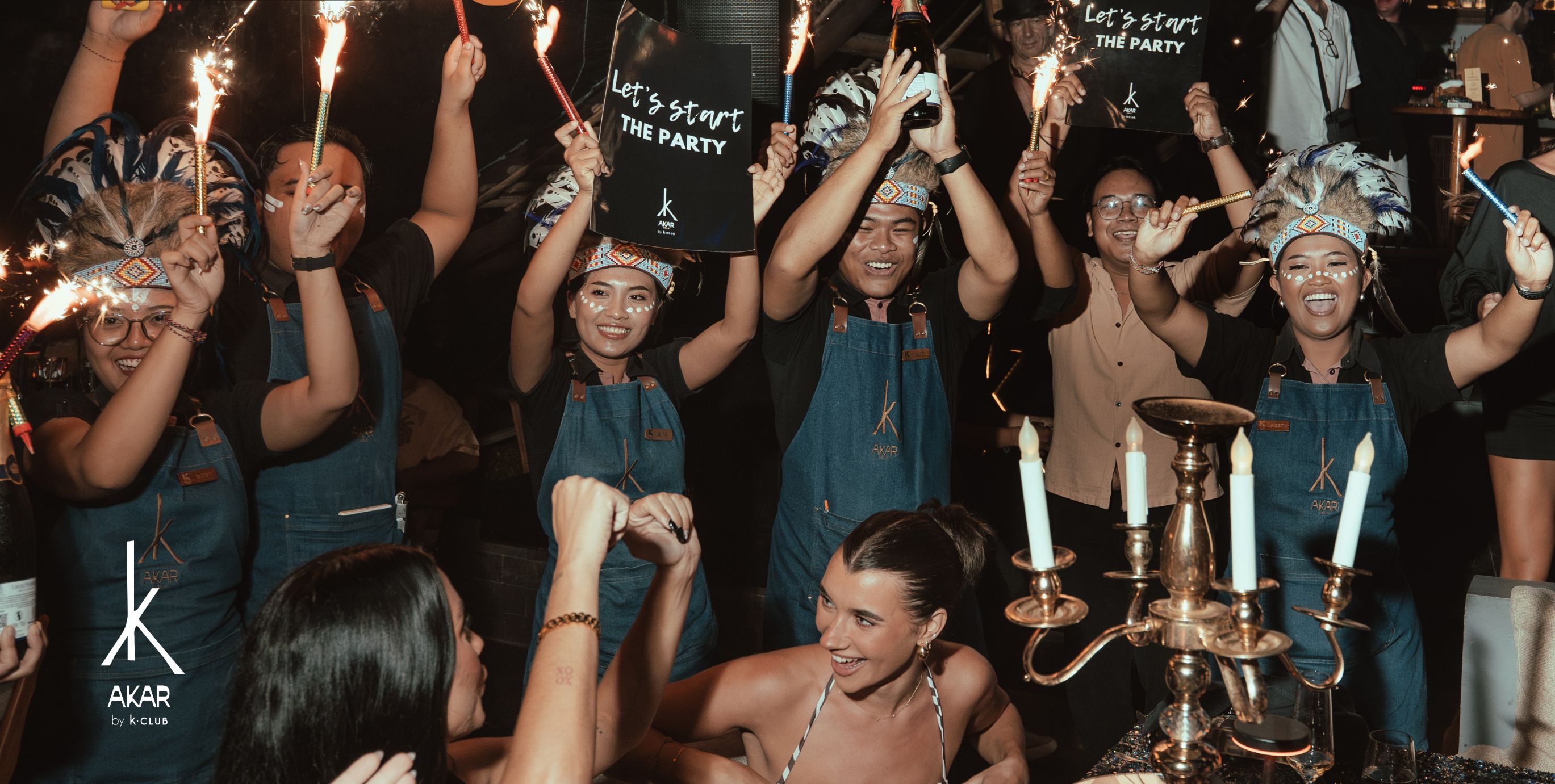 Un groupe de personnes fait la fête dans un restaurant gastronomique de luxe, et une pancarte indique : « Que la fête commence ! »