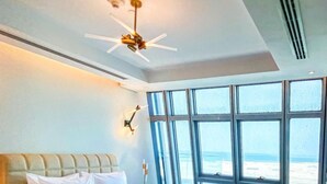 A hotel room with a large bed, a ceiling fan, a wall-mounted light, and a view of the ocean through large windows. - WATERFRONT HOTEL  APARTMENT (Lusail)