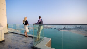A high-rise building with a glass balcony overlooking a body of water and a cityscape. - WATERFRONT HOTEL  APARTMENT (Lusail)