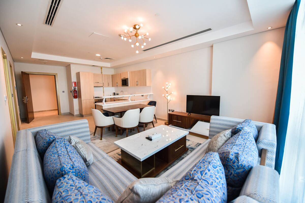 A modern living room with a sofa set, a coffee table, and a TV. There is a kitchen area with cabinets and a microwave.