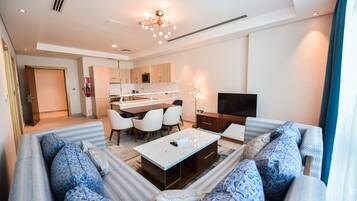 A modern living room with a sofa set, a coffee table, and a TV. There is a kitchen area with cabinets and a microwave.