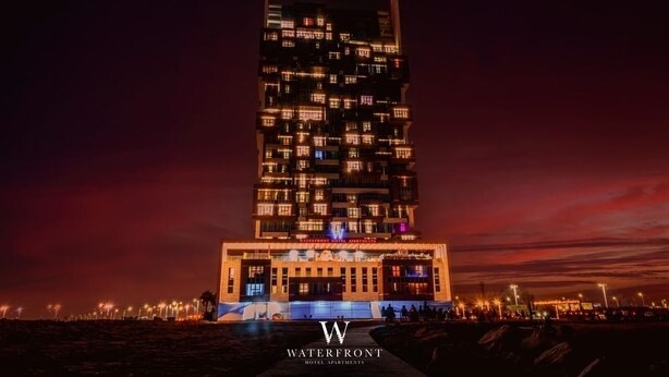 A modern hotel with a unique, multi-story design, featuring numerous balconies and a prominent entrance area. - WATERFRONT HOTEL  APARTMENT (Lusail)