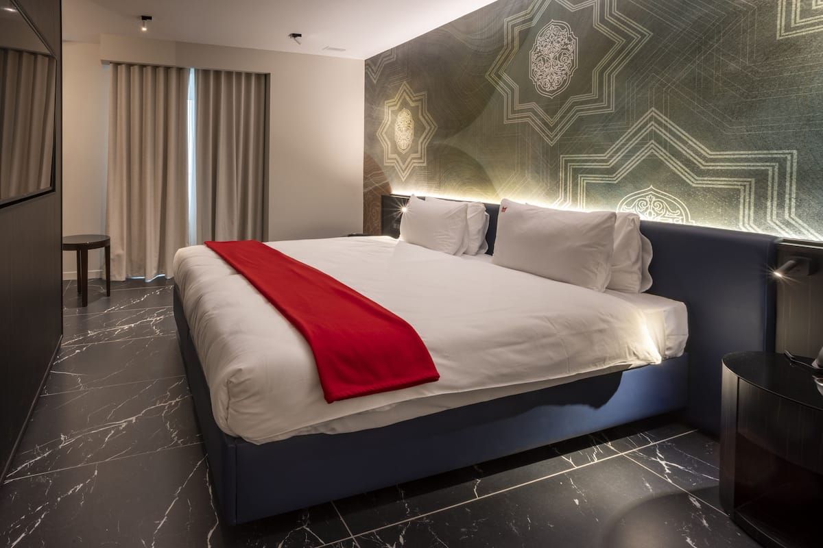 A modern hotel room with a large bed, a red throw blanket, a headboard with built-in lighting, a marble floor, and a geometric-patterned wall.