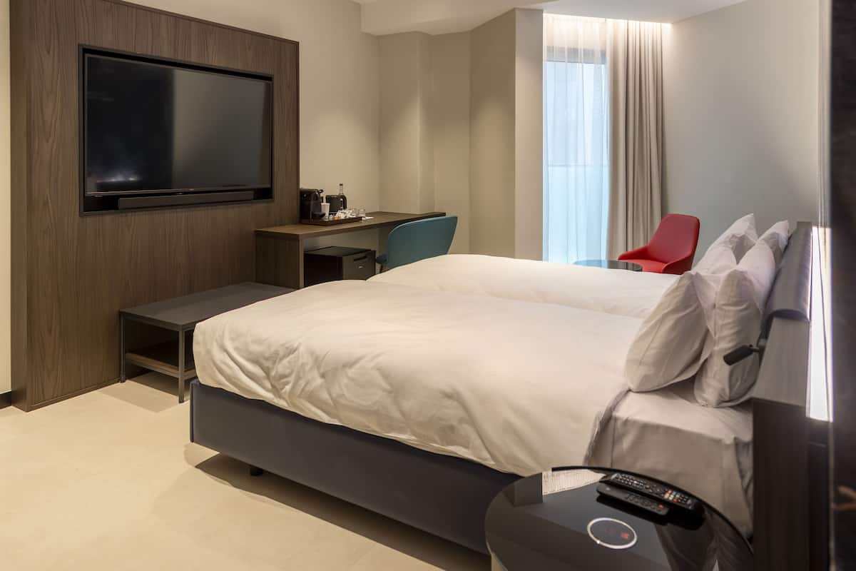 A modern hotel room with a large bed, a desk with a chair, a television mounted on the wall, and a window with curtains.