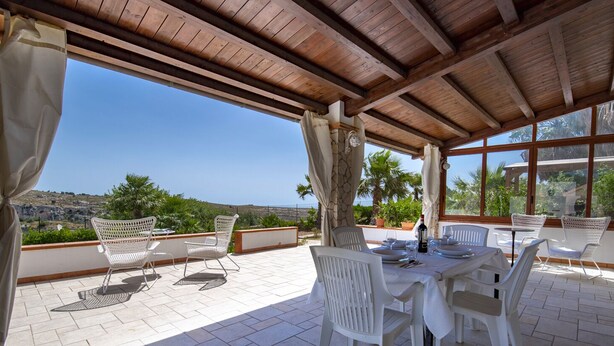 Un patio al aire libre con sillas y mesas de mimbre blanco, techo de madera y vista al paisaje.
