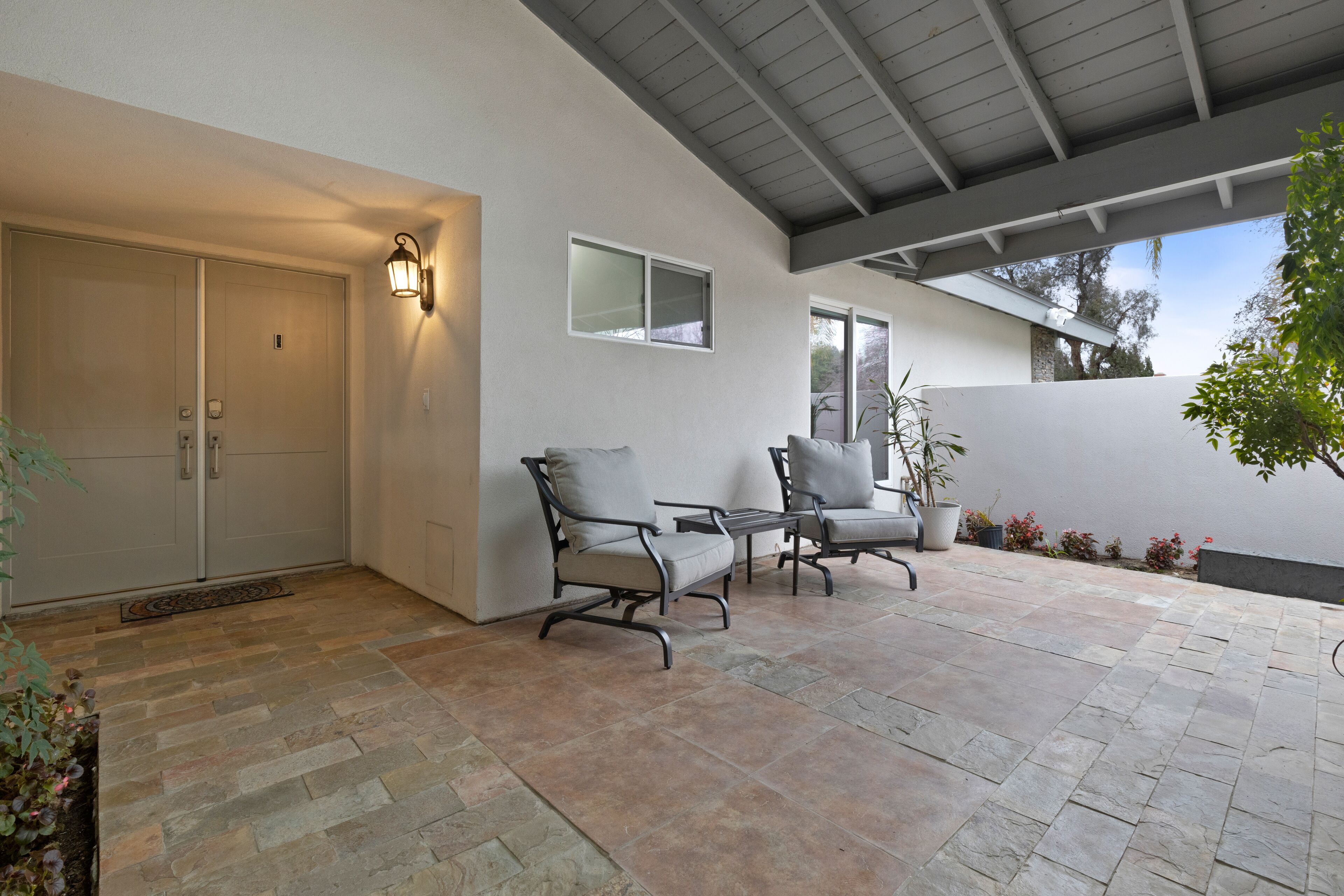 Eine überdachte Terrasse mit Steinboden, zwei gepolsterte Stühle, ein kleiner Tisch und eine Wandleuchte.