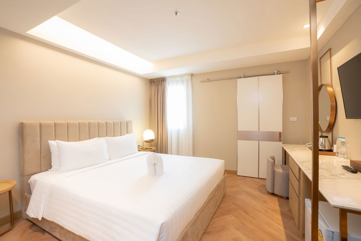 Habitación de hotel con una cama grande, una mesita de noche con lámpara, un armario, un tocador con espejo y un televisor.