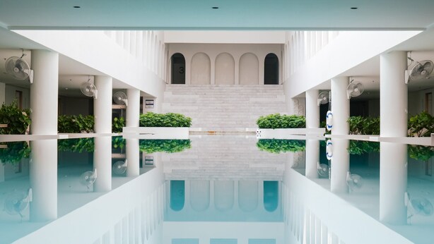 En moderne hotellobby med en central pool, hvide søjler og buede døråbninger.