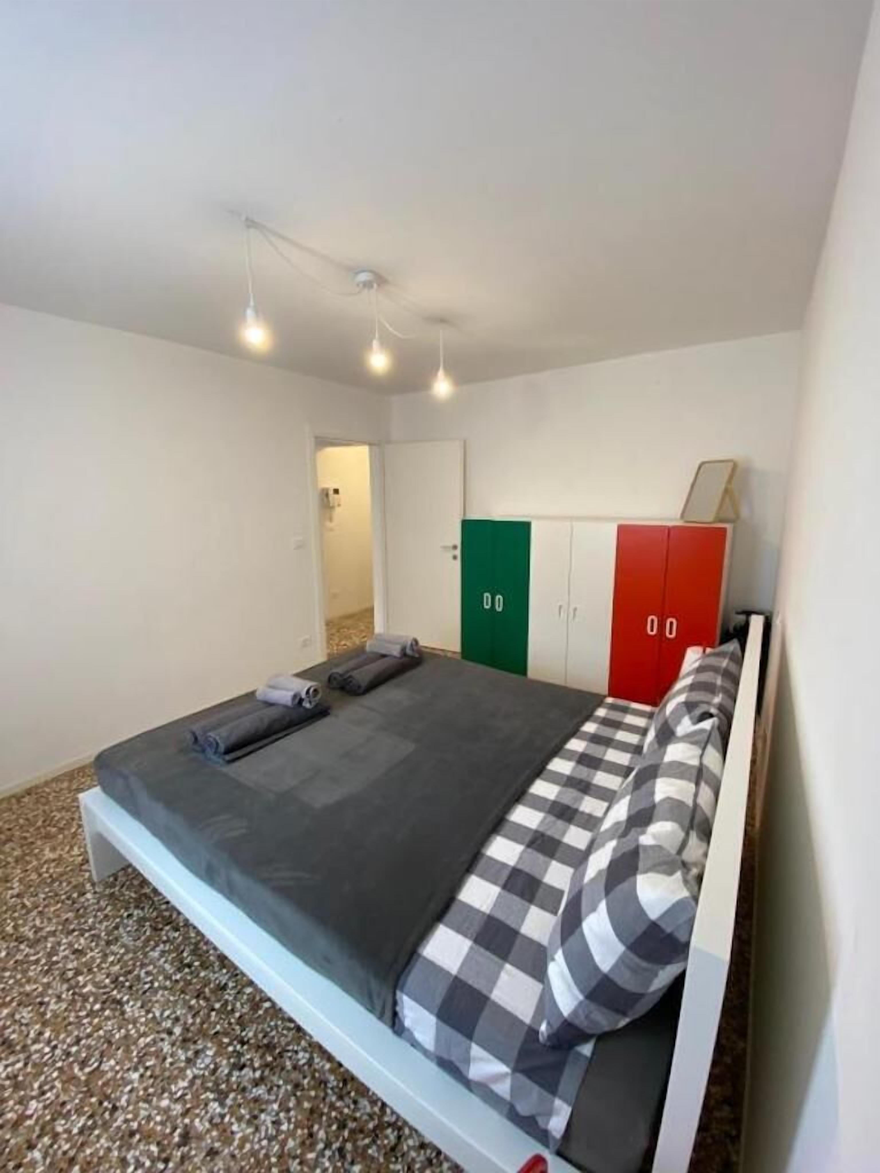 Un letto singolo con coperta e cuscini a quadri, un comodino, un armadio con ante verdi e rosse e un lampadario a soffitto.