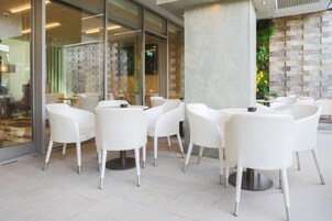 Un área de estar interior moderna con sillas y mesas blancas, un muro de vidrio y una pared texturizada con diseño de ladrillo.