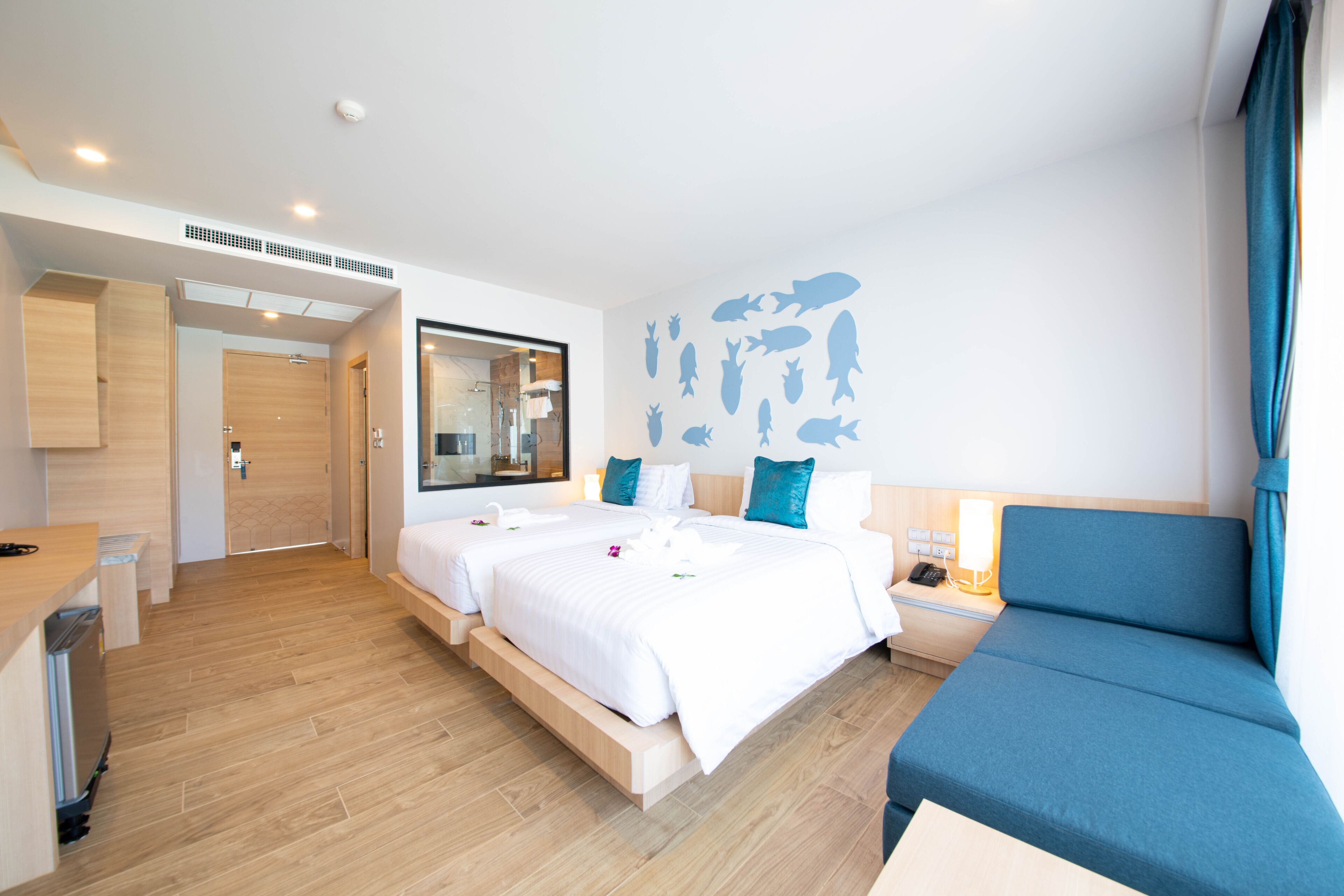 A hotel room with two beds, a desk, a mirror, a wall with fish silhouettes, and a blue sofa.