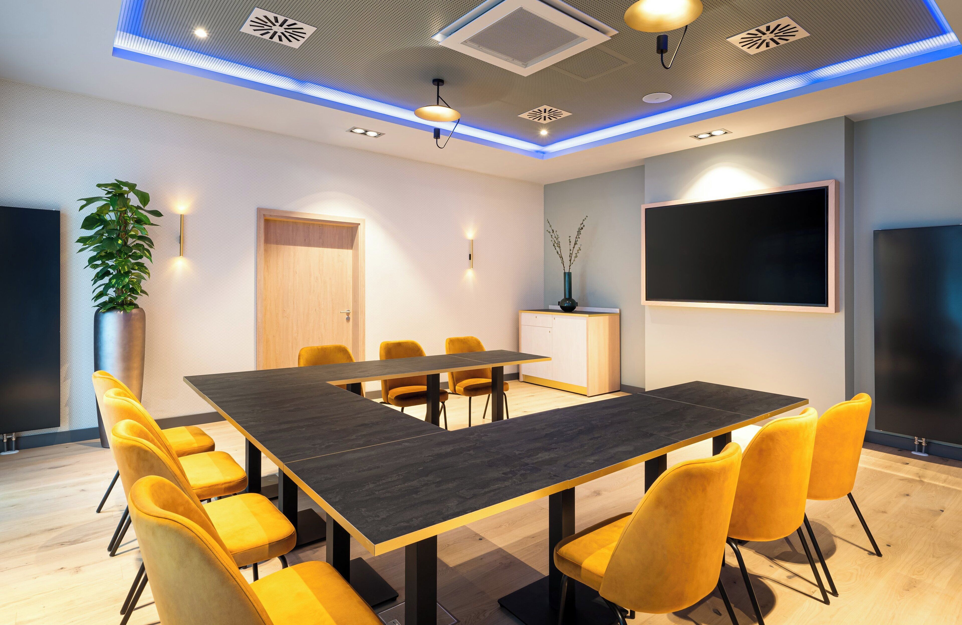 A conference room with a long table, yellow chairs, a potted plant, a cabinet, and a large screen.