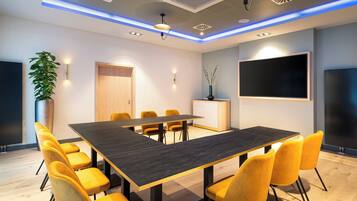 A conference room with a long table, yellow chairs, a potted plant, a cabinet, and a large screen.